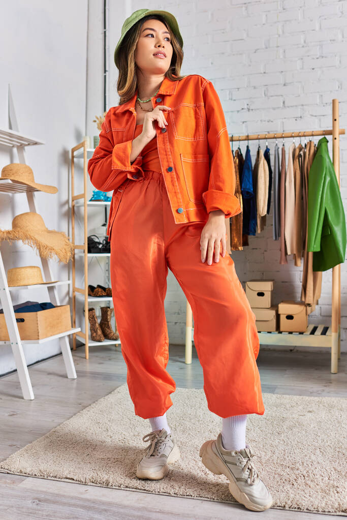 pewny azjatycki projektant w pomarańczowy odzież i kapelusz panama pozowanie w nowoczesny osobisty atelier - Zdjęcie, obraz