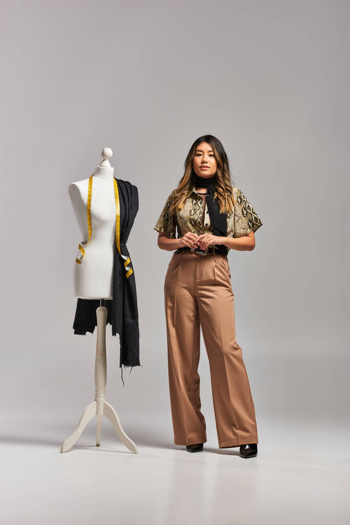 moda asiático diseñador de moda posando cerca maniquí con cinta métrica y tela sobre fondo gris - Foto, imagen