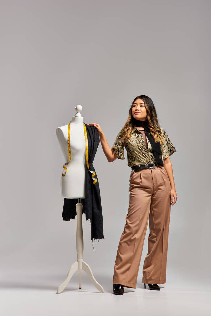 успешный модный азиатский дизайнер, смотрящий вблизи манекена с тканью и измерительной лентой на сером - Фото, изображение
