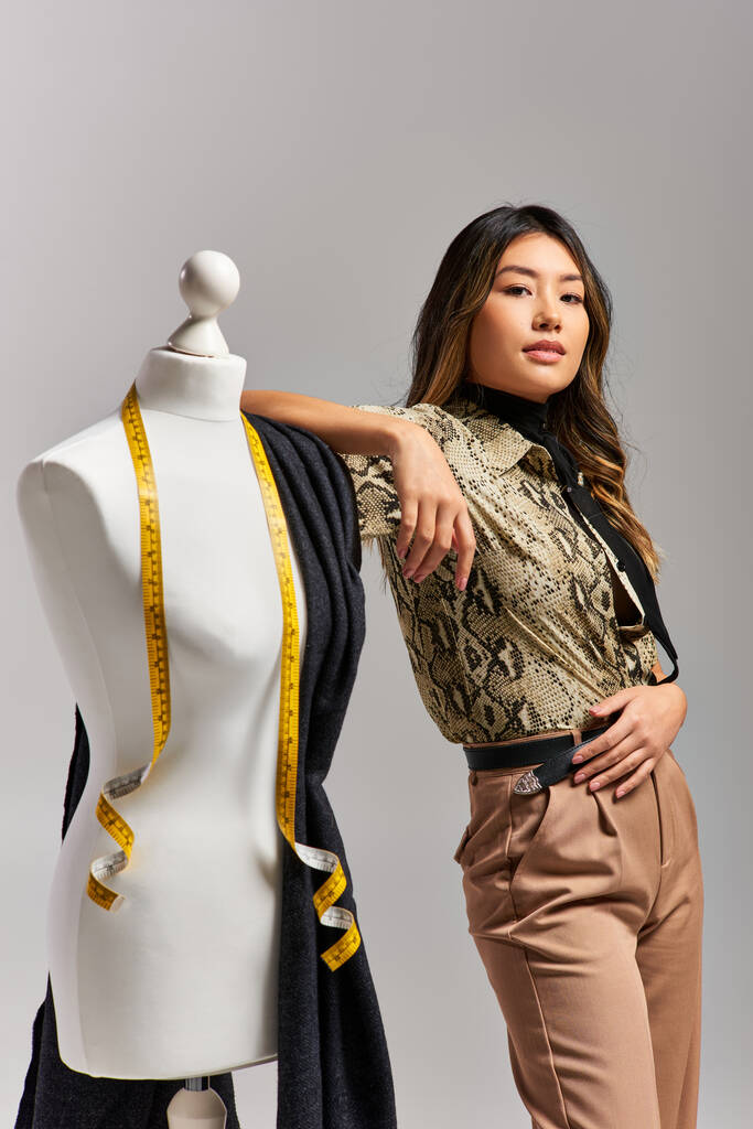 Genç ve çekici Asyalı tasarımcı kumaş ve mezura ile mankenin yakınındaki kameraya bakıyor. - Fotoğraf, Görsel
