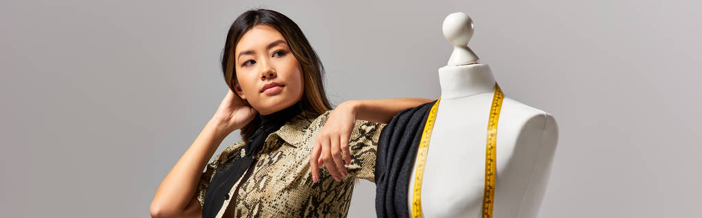 Modern Asyalı stilist kumaşla mankenin yanından uzağa bakıyor ve gri bantla ölçüm yapıyor. - Fotoğraf, Görsel