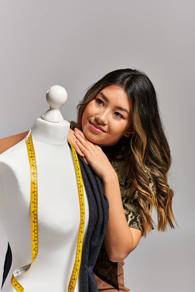 Hayalperest Asyalı kıyafet tasarımcısı gri arkaplanda şerit ile mankenin yanından uzağa bakıyor. - Fotoğraf, Görsel
