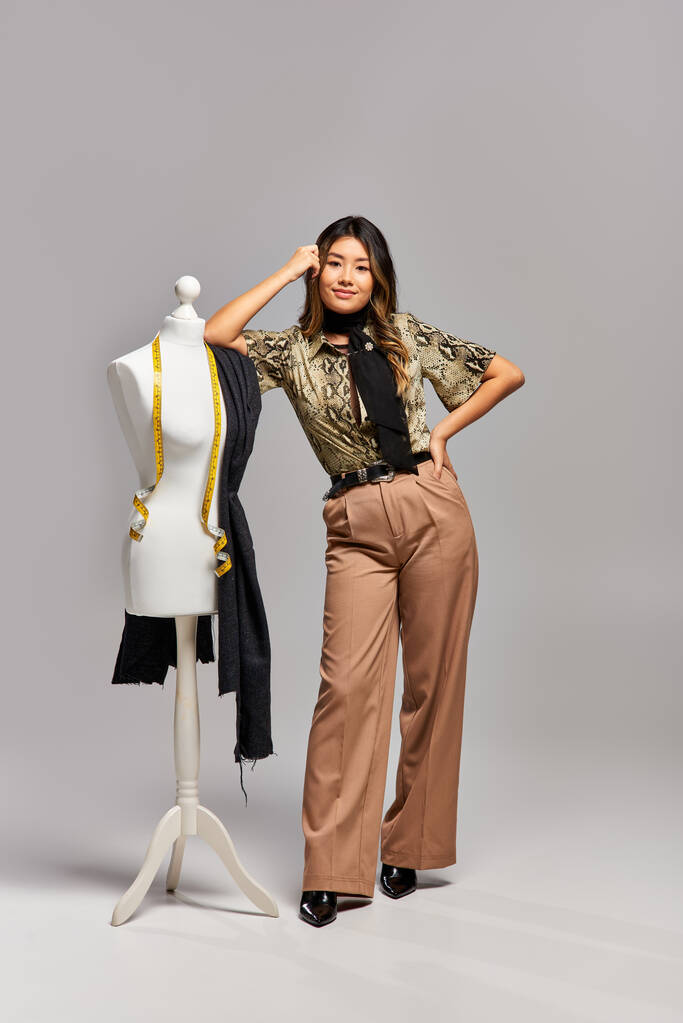 trendy asian fashion designer posing near mannequin with measuring tape and fabric on grey backdrop - Photo, Image