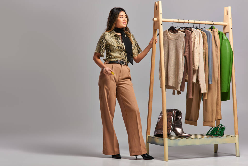 счастливый азиатский дизайнер с измерительной лентой глядя в сторону стойки со стильной одеждой на сером - Фото, изображение