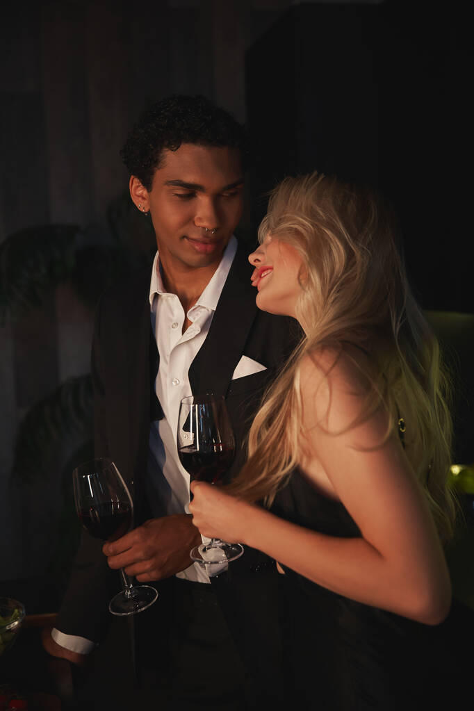 веселая молодая мультикультурная пара в элегантных нарядах проводит время вместе и пьет красное вино - Фото, изображение