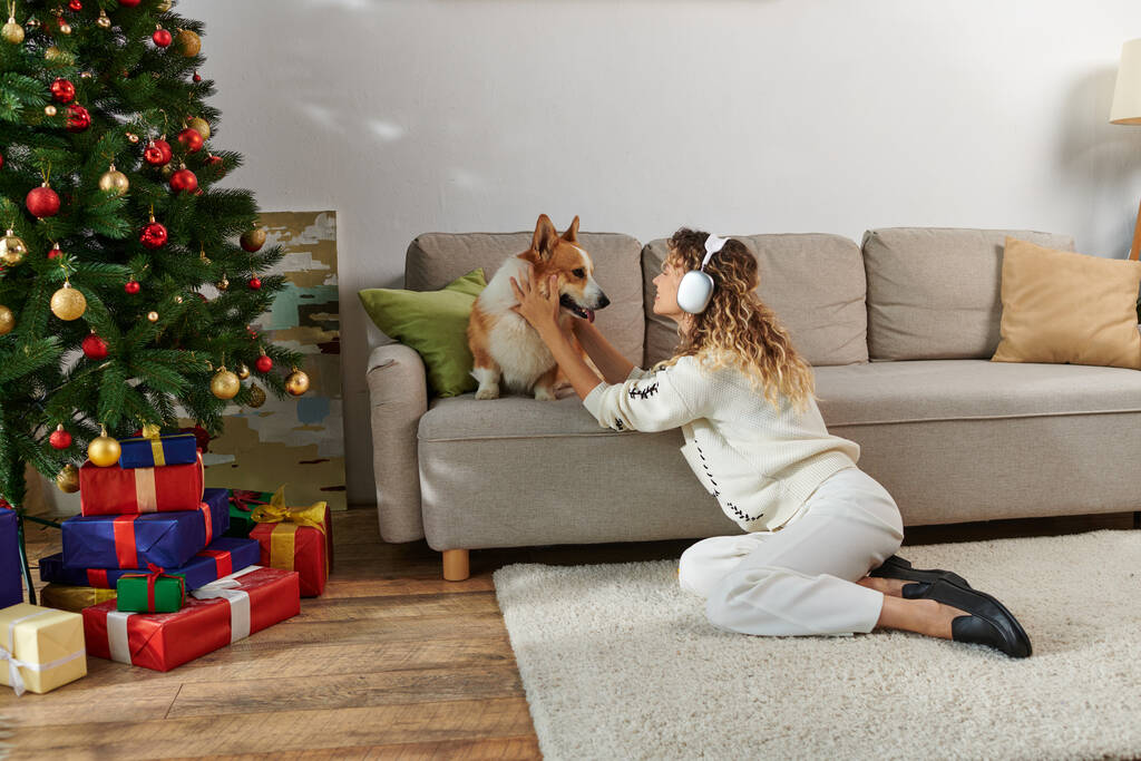 curly woman in wireless headphones playing with cute corgi dog near decorated Christmas tree - Photo, Image