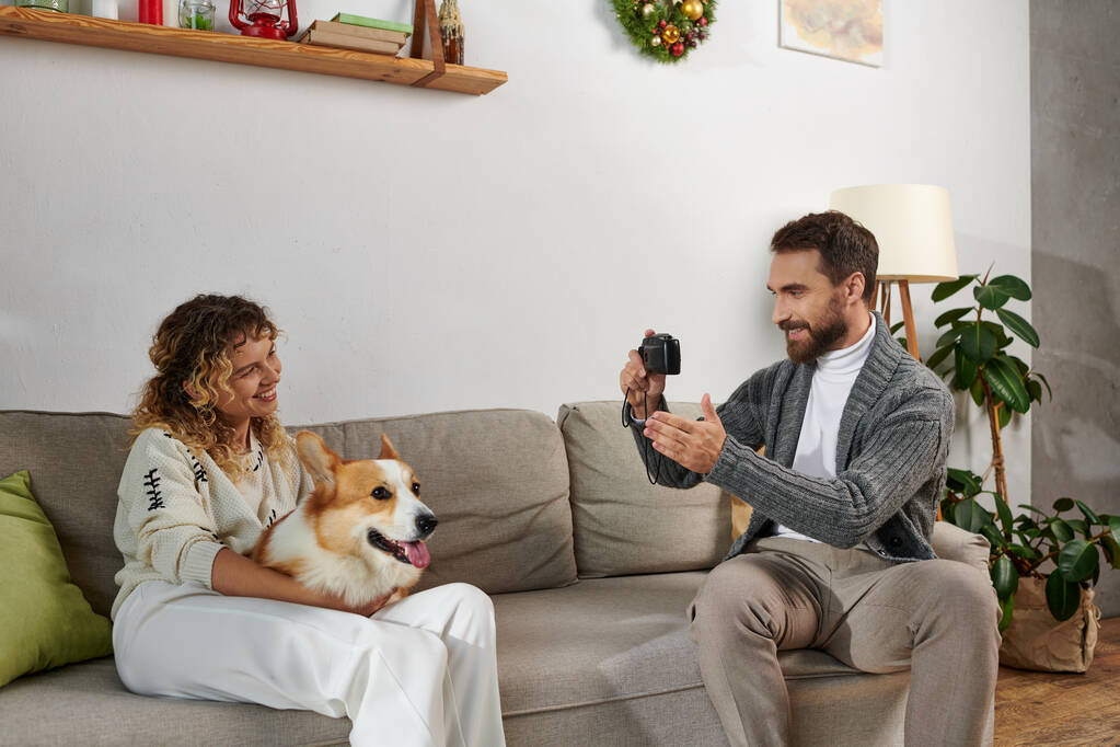 Mann lächelt und fotografiert Frau mit Corgi-Hund in moderner Wohnung, glückliche Momente - Foto, Bild