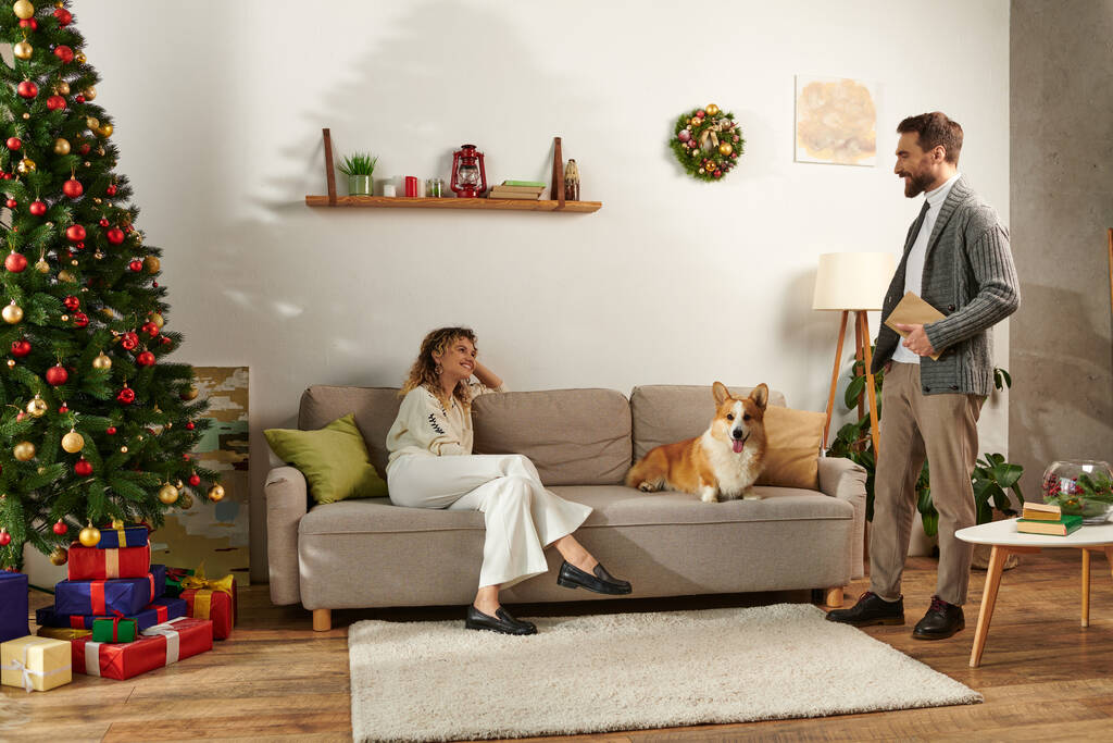 femme heureuse assise sur le canapé avec chien corgi et regardant mari avec livre le jour de Noël - Photo, image