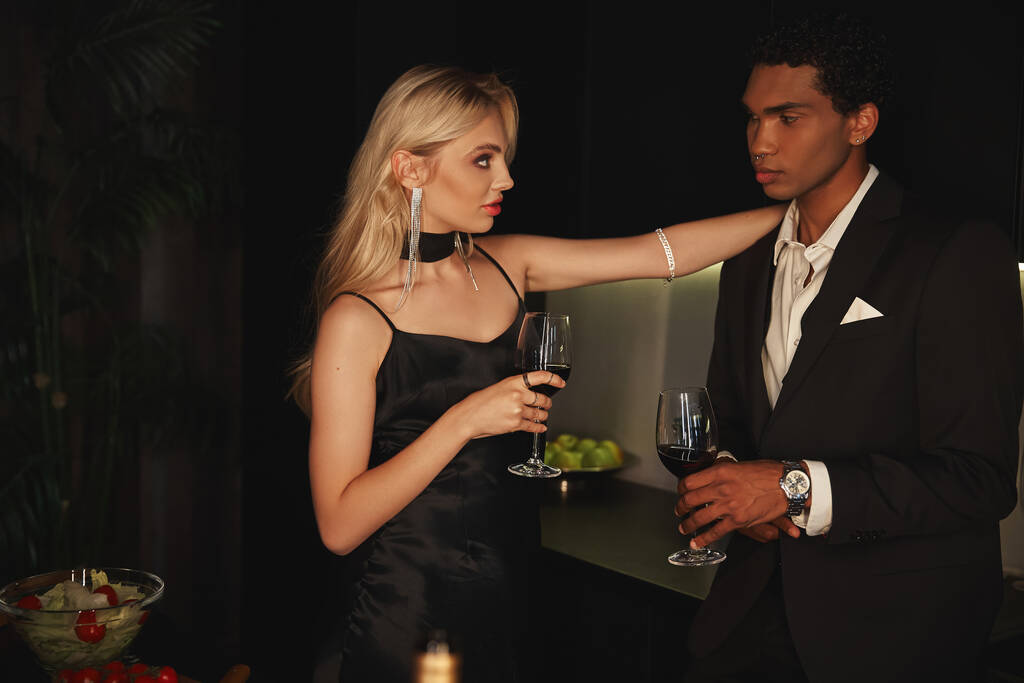 atractiva pareja sensual diversa en elegantes trajes sosteniendo copas de vino y mirándose - Foto, Imagen