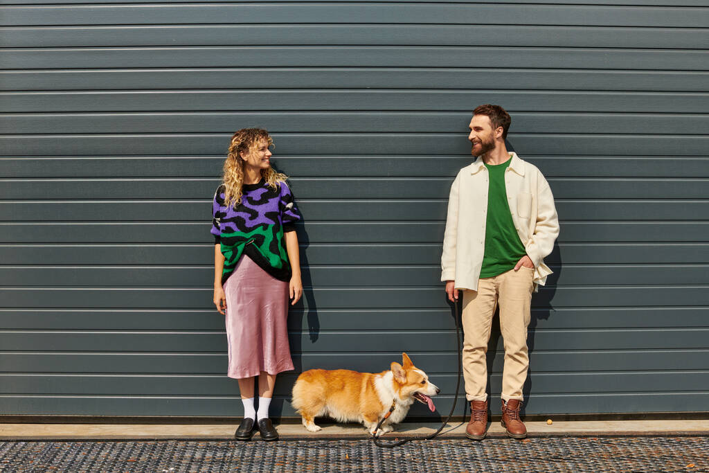 glückliches und stilvolles Paar beim Gassigehen mit Corgi-Hund in der Nähe von grauen Garagentoren, tierischen Begleitern - Foto, Bild