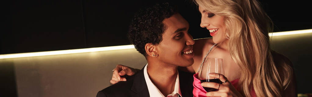 giovane donna che tiene un bicchiere di vino rosso e sorride gioiosamente al suo ragazzo afroamericano, striscione - Foto, immagini