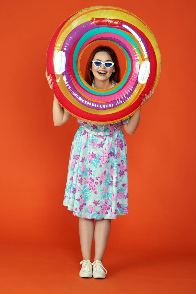 Reizen en vakantie reclame concept. Studio shot van vrouwelijk model. Vrouw voor de camera kijkend door kleurrijke opblaasbare buis glimlachend, geïsoleerd op oranje achtergrond. - Foto, afbeelding