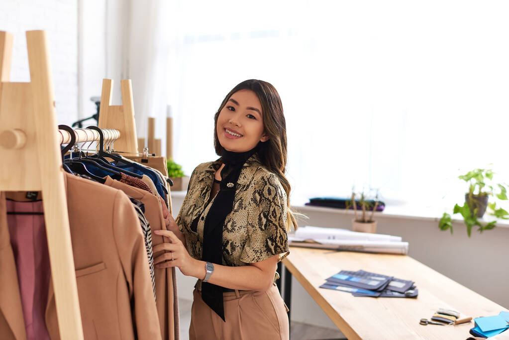 vrolijk aziatische mode stylist kijken naar camera in de buurt rack met op maat gemaakte kleding in prive-atelier - Foto, afbeelding