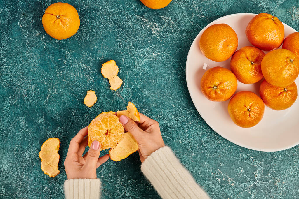 částečný pohled na ženu loupající čerstvě zralé mandarinky na modrém texturovaném povrchu, vánoční koncept - Fotografie, Obrázek