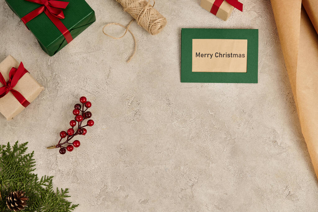 Feliz Natal cartão perto de caixas de presente e ramos de zimbro com bagas de azevinho em cinza - Foto, Imagem