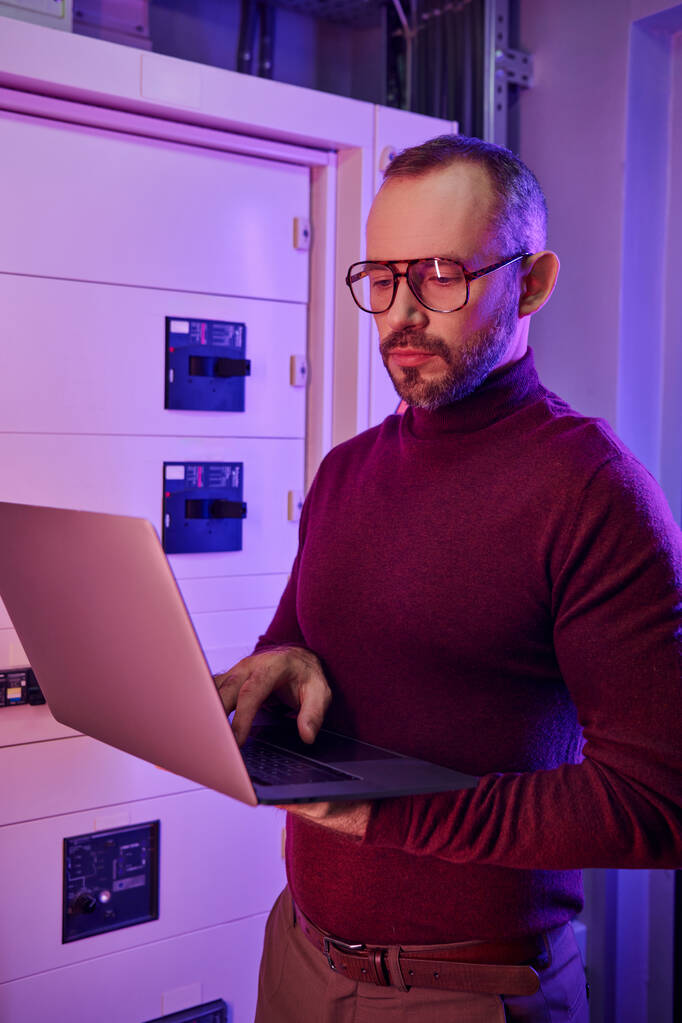attraente specialista focalizzato in dolcevita con barba e occhiali guardando attentamente il suo computer portatile - Foto, immagini