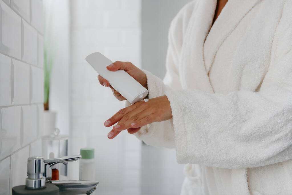 Крупным планом женщина в халате наносит косметический крем на руку, стоя в ванной комнате - Фото, изображение