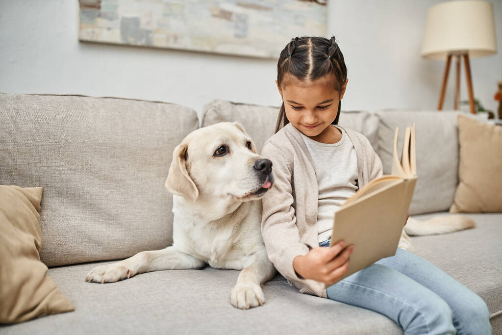 szczęśliwa dziewczyna siedzi na kanapie i czytania książki w pobliżu labrador w nowoczesnym salonie, towarzysz zwierząt - Zdjęcie, obraz