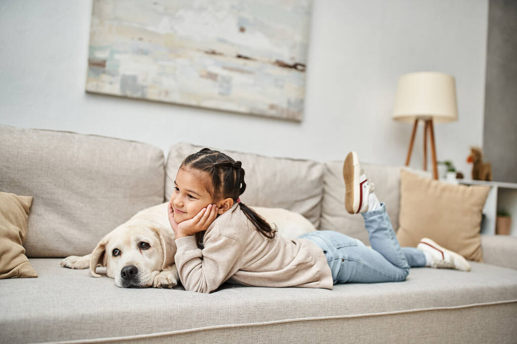 gelukkig meisje glimlachen en liggen op de bank met schattige labrador in moderne woonkamer, huisdier en kind - Foto, afbeelding