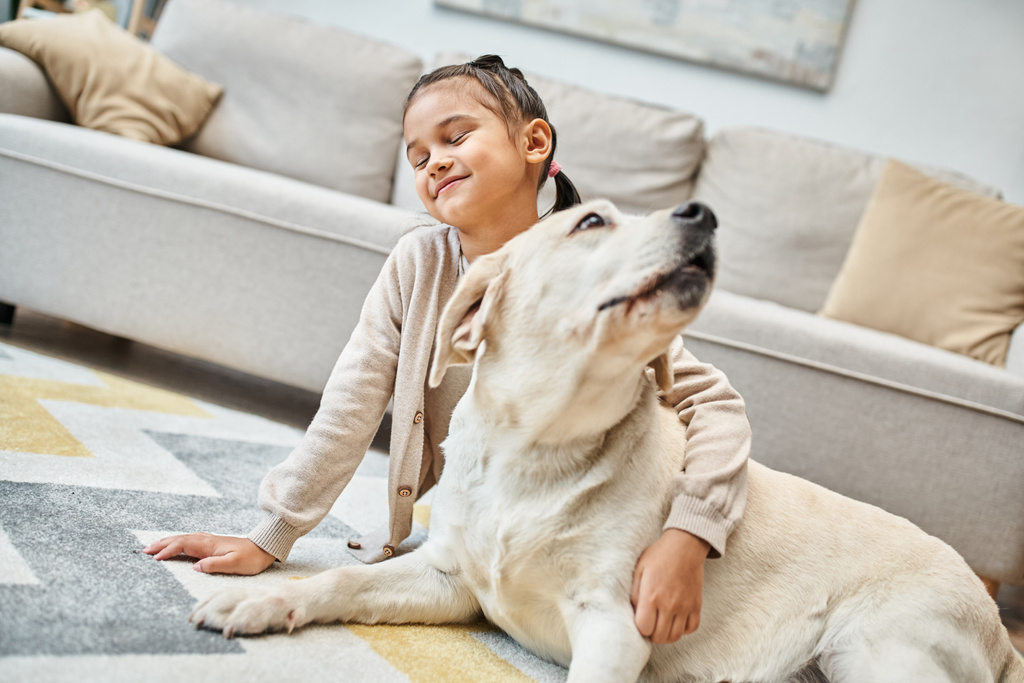 zadowolony dziewczyna siedzi na dywanie i głaskanie labrador w nowoczesnym salonie, dziecko i futrzany przyjaciel - Zdjęcie, obraz