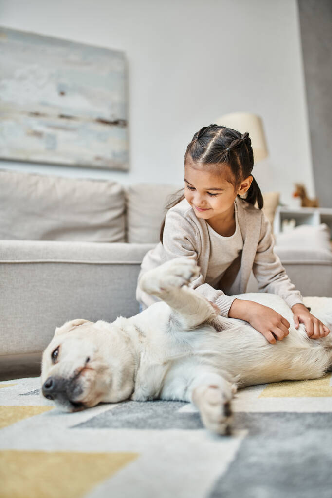 позитивная девушка в повседневной одежде улыбается и гладит собаку в современной гостиной, ребенок и лабрадор - Фото, изображение