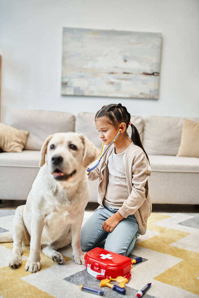chica feliz en traje casual jugando médico con perro labrador en la sala de estar, kit de primeros auxilios de juguete - Foto, Imagen