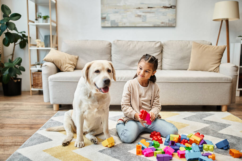 fröhliches Mädchen spielt mit bunten Spielzeugklötzen in der Nähe Labrador im Wohnzimmer, Gebäude Turm Spiel - Foto, Bild