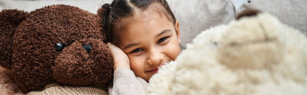 alegre chica de edad elemental sentado entre suaves osos de peluche en el sofá en la sala de estar moderna, pancarta - Foto, Imagen
