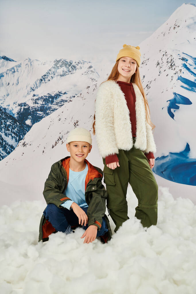 függőleges lövés a kisfiú ül a hóban közel aranyos álló lány, stílusos ruha, téli divat - Fotó, kép