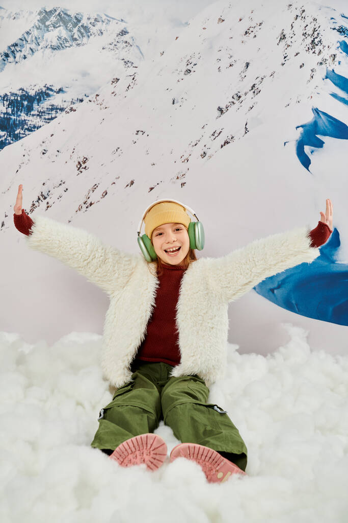 tiro vertical de chica en traje cálido con auriculares sentados en la nieve animando con los brazos levantados - Foto, Imagen