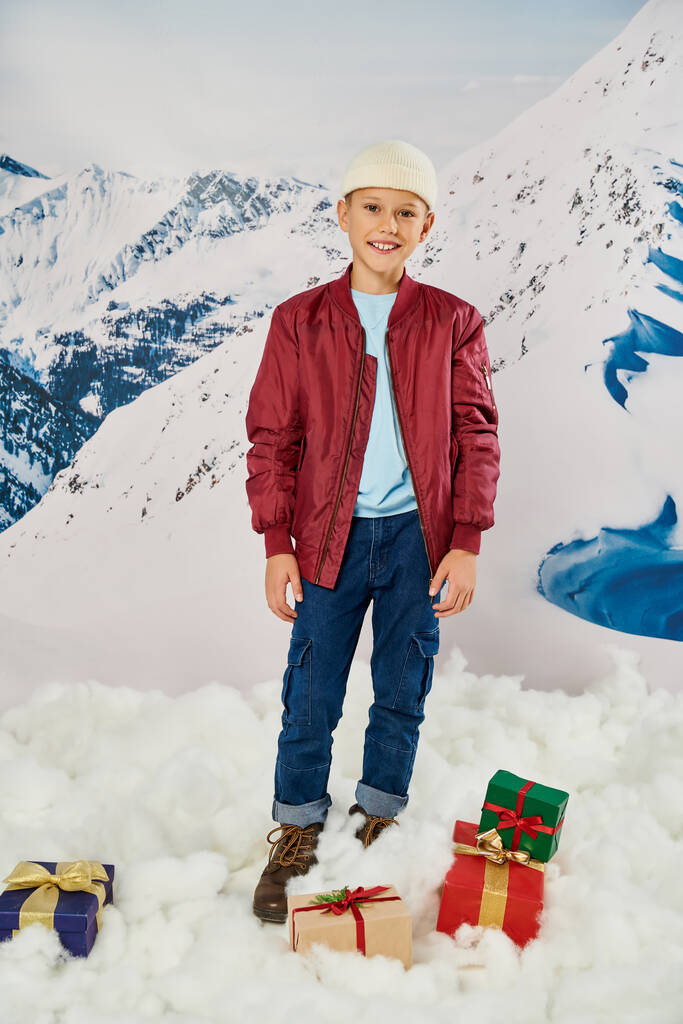 tiro vertical de menino pré-adolescente em casaco elegante vermelho posando ao lado de presentes e sorrindo para a câmera - Foto, Imagem