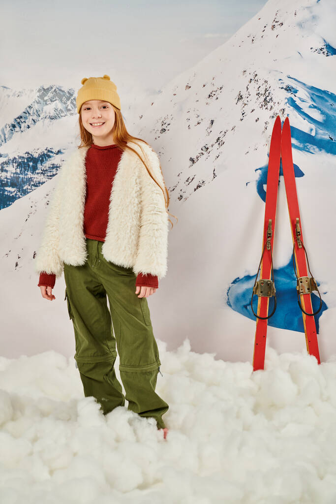 verticaal schot van vrolijke tiener meisje in trendy warme kleding poseren met ski 's op de achtergrond, mode - Foto, afbeelding