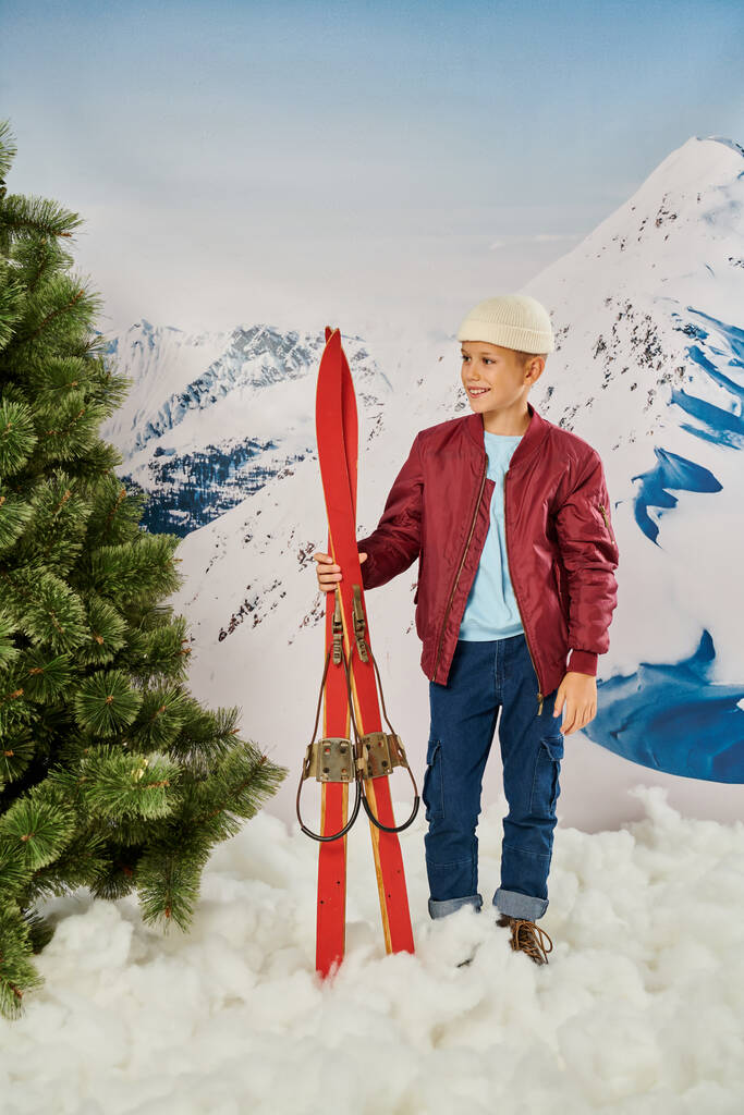 вертикальний знімок маленького стильного хлопчика в теплому вбранні з лижами, радісно посміхаючись, концепція моди - Фото, зображення