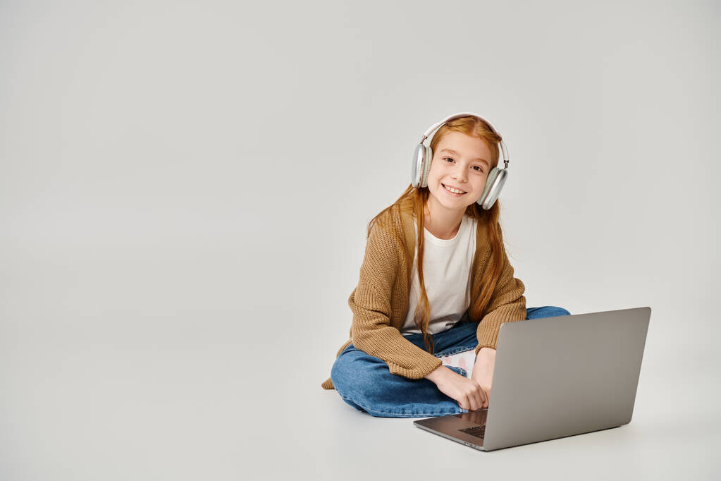 vrolijk pre-adolescent meisje op de vloer met laptop en headset op grijze achtergrond, mode concept - Foto, afbeelding