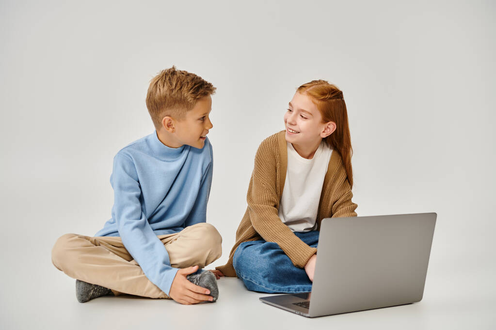 радісні передпідлітки діти сидять на підлозі з ноутбуком і посміхаються один одному, мода - Фото, зображення