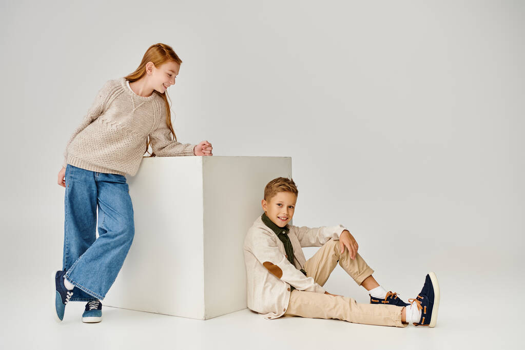 wesoła nastolatka dziewczyna stoi obok biały sześcian i patrząc na małego chłopca siedzi na podłodze, moda - Zdjęcie, obraz