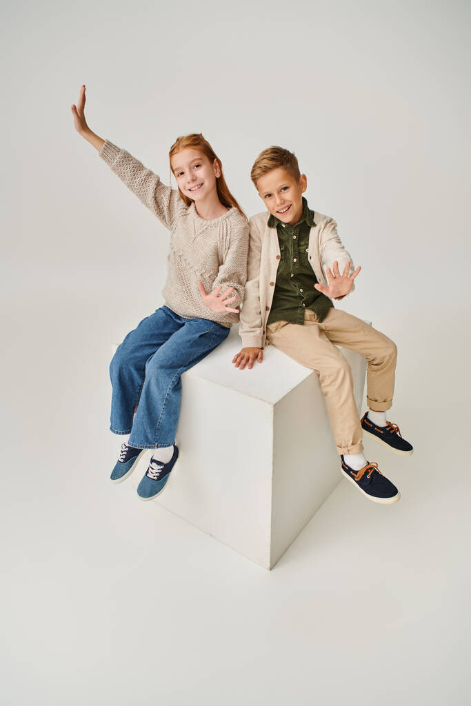 κάθετη λήψη χαρούμενων μικρών φίλων με χειμωνιάτικα ρούχα να κάθονται σε λευκό κύβο και να χαιρετάνε την κάμερα - Φωτογραφία, εικόνα