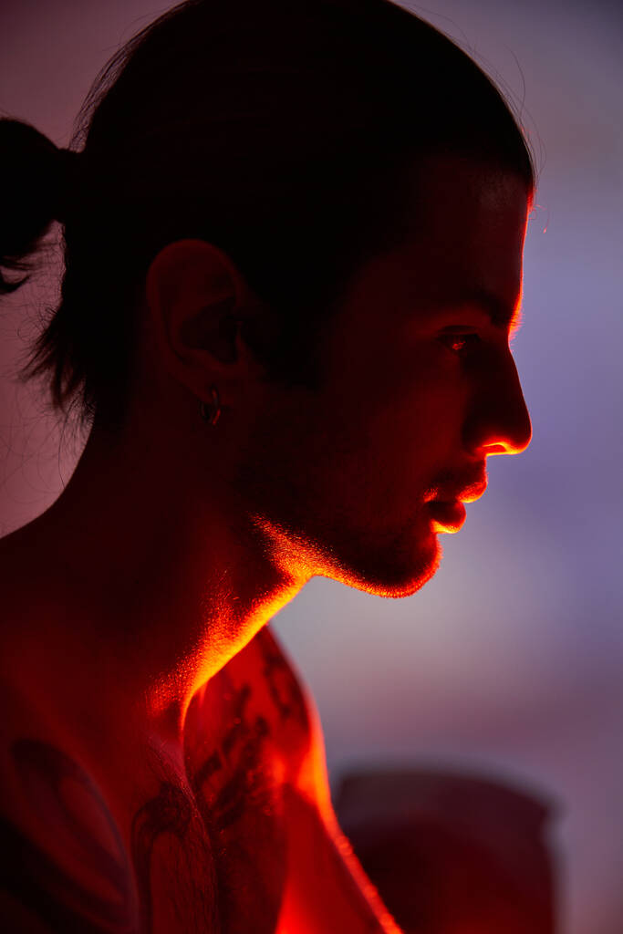 joven hombre guapo con tatuajes en el cuerpo y pendientes posando en perfil rodeado de vibrantes luces - Foto, Imagen