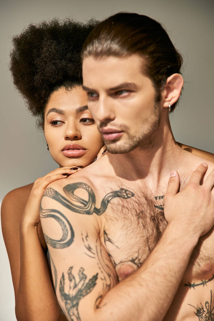 csábító férfi tetoválásokkal ölelgeti a csinos afro-amerikai barátnőjét szürke háttérben. - Fotó, kép