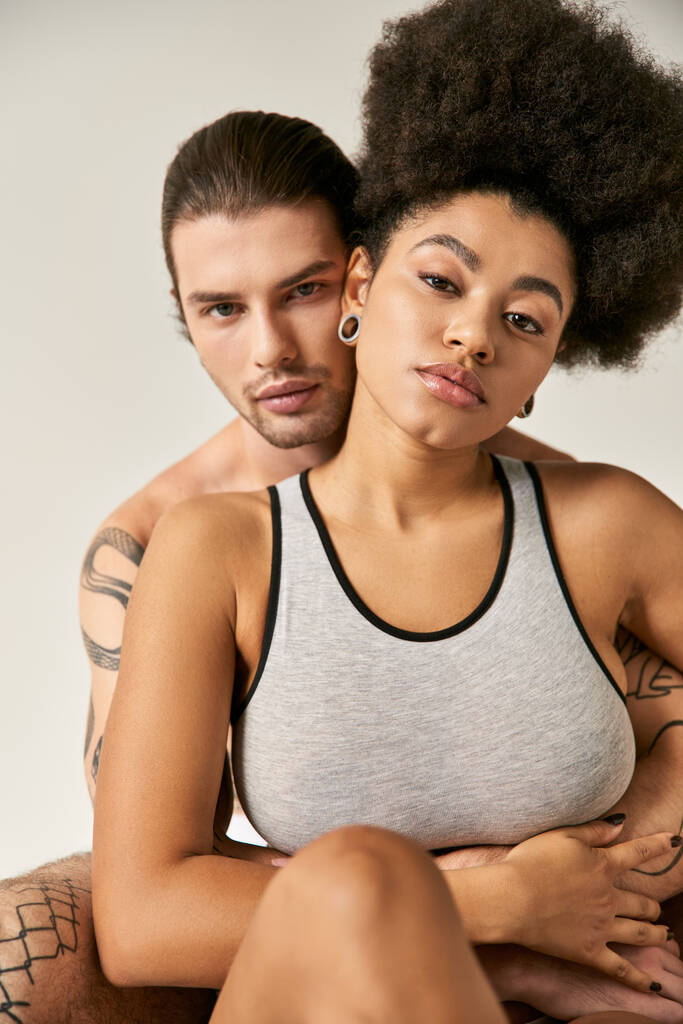 young tattooed man hugging his attractive african american girlfriend on gray backdrop, sexy couple - Photo, Image