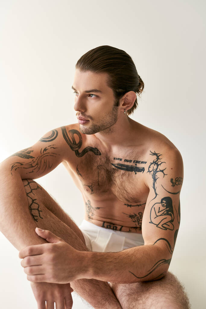 hombre joven y guapo tatuado con cola de caballo en ropa interior sentado en el suelo y mirando hacia otro lado - Foto, imagen
