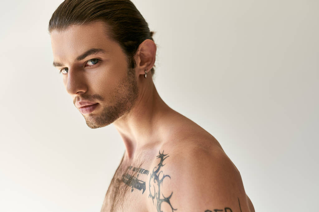 uwodzicielski młody mężczyzna z kucykiem i fajnymi tatuażami w wygodnej bieliźnie pozujący na tle ecru - Zdjęcie, obraz