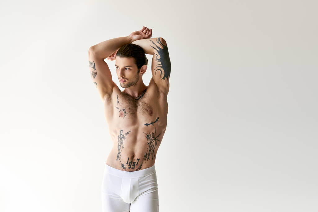 sexy verführerischer Mann mit Pferdeschwanz und coolen Tattoos in bequemer Unterwäsche posiert auf ecru Hintergrund - Foto, Bild