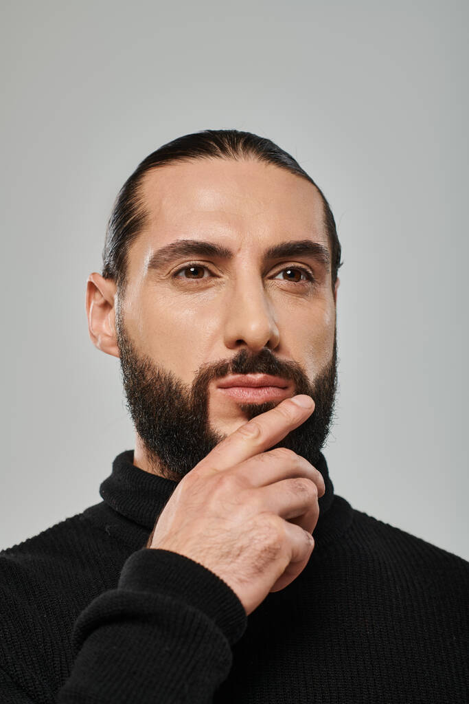 portrait of good looking arabic man in turtleneck touching beard while thinking on grey backdrop - Photo, Image