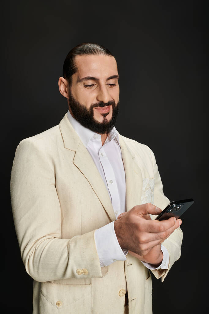 黒い背景にスマートフォンを使用して白いシャツとブレーザーで陽気でひげ付きのアラビア人男性 - 写真・画像