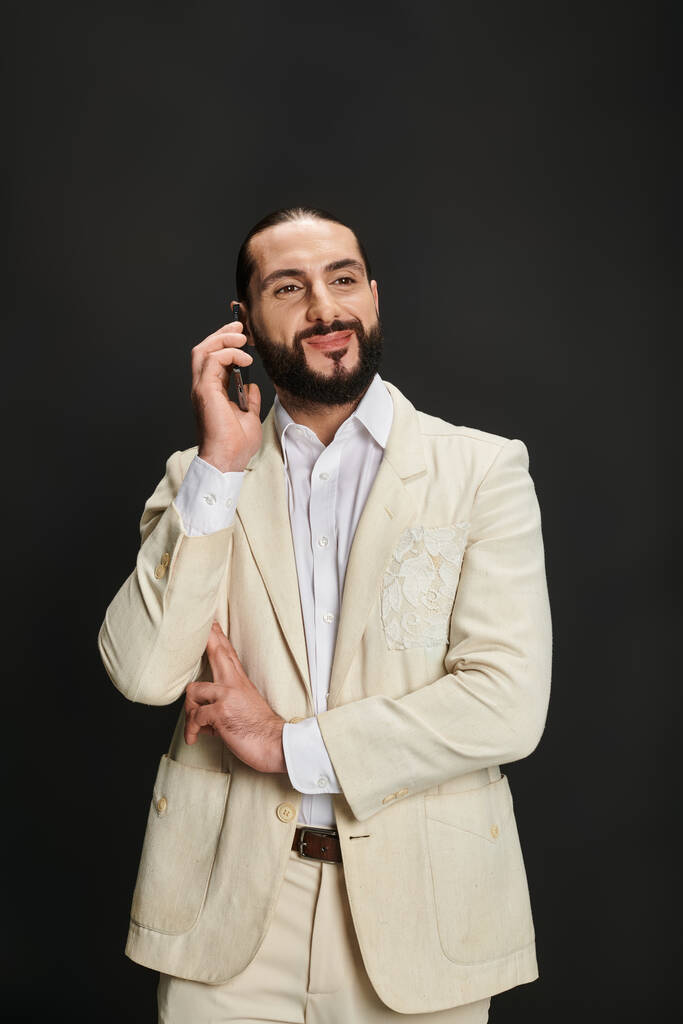 Beyaz gömlekli ve ceketli neşeli ve sakallı Arap adam siyah arka planda akıllı telefondan konuşuyor. - Fotoğraf, Görsel
