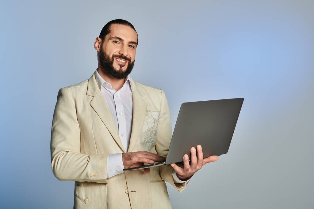 hombre árabe barbudo en ropa formal elegante usando portátil sobre fondo gris, era digital - Foto, Imagen