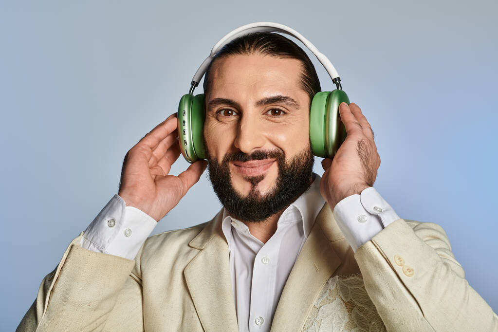 felice uomo barbuto in elegante abbigliamento formale ascoltare musica in cuffie wireless su sfondo grigio - Foto, immagini