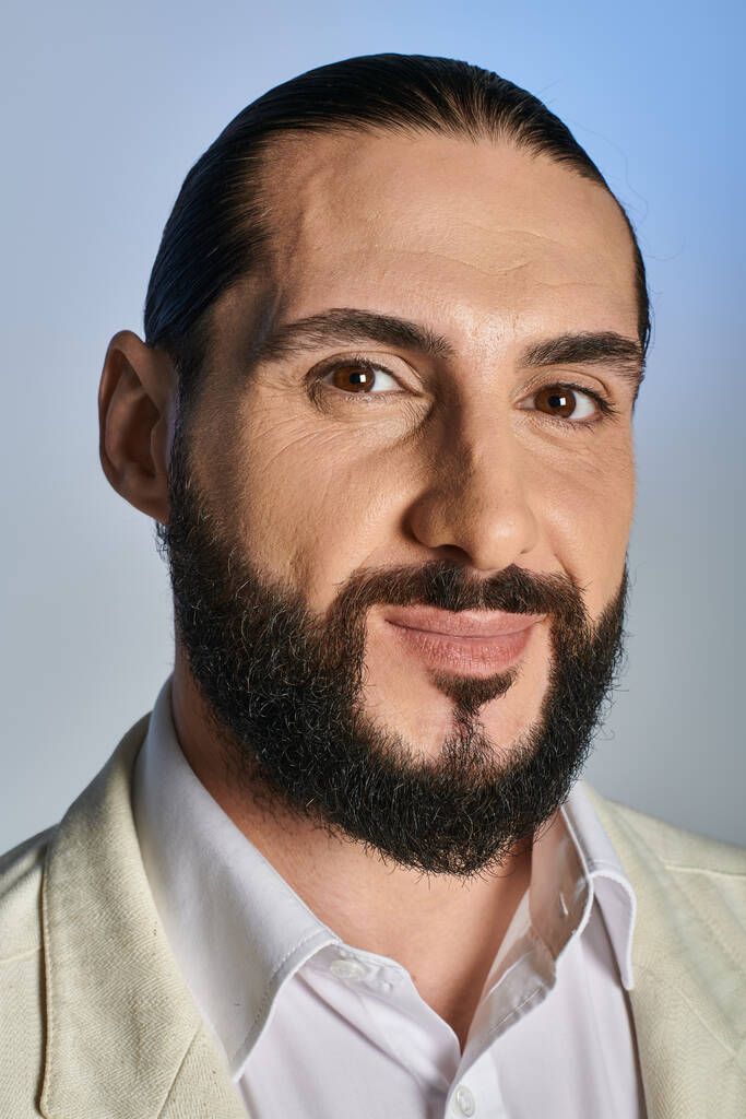 Porträt eines gutaussehenden und bärtigen arabischen Mannes in weißer eleganter Kleidung, der lächelt und die Kamera auf grau legt - Foto, Bild