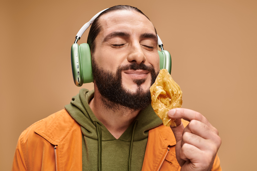 щасливий арабський чоловік в навушниках пахне медовою баклавою на бежевому фоні, середній східний десерт - Фото, зображення